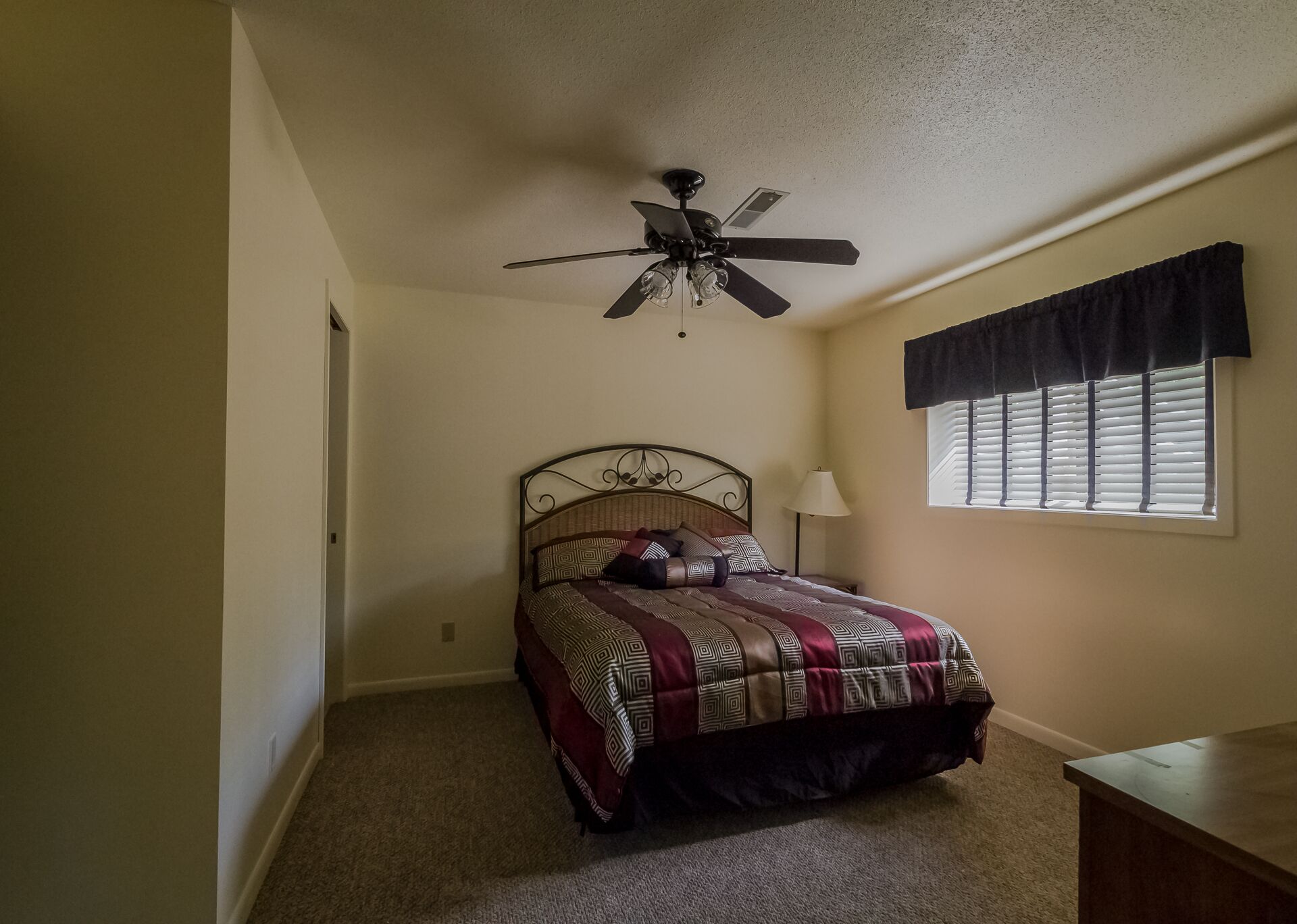 A bedroom with a bed, a ceiling fan, a window with blinds, and a nightstand with a lamp.
