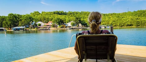 A person sitting on a wooden deck chair overlooking a lake with houses and boats in the background.