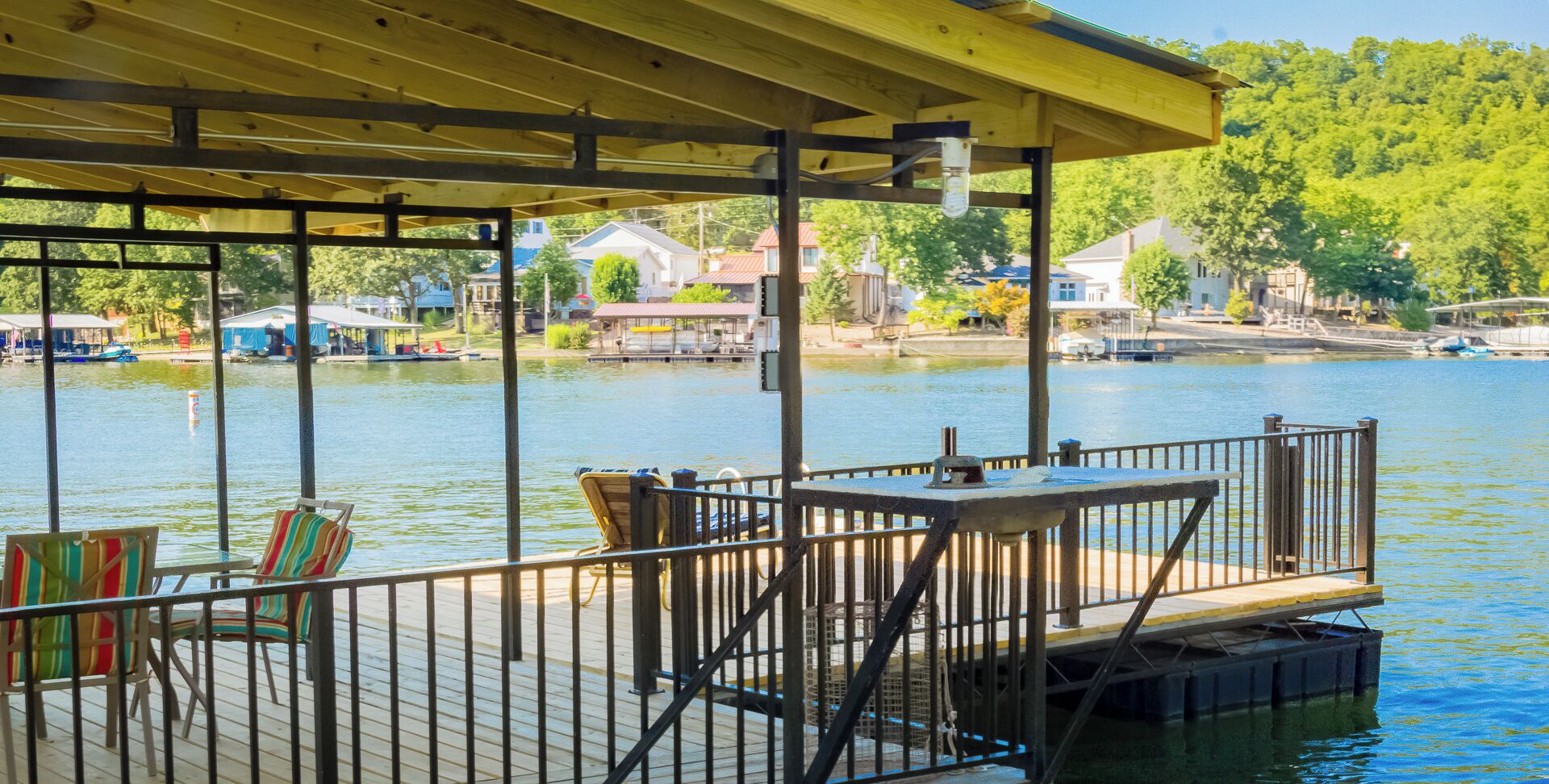 Une terrasse en bois, agrémentée de chaises et d’une table, offre une vue sur un lac paisible, entouré de maisons et de bateaux.