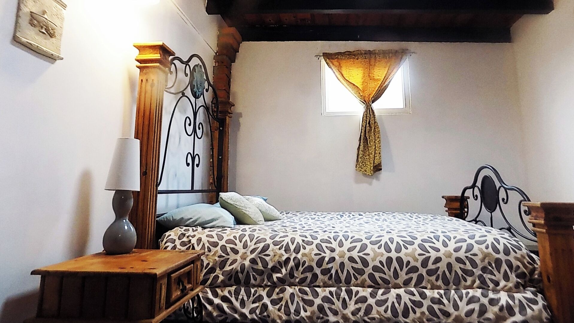 Un dormitorio con cama, mesita de noche, lámpara, ventana con cortina y estante en la pared.