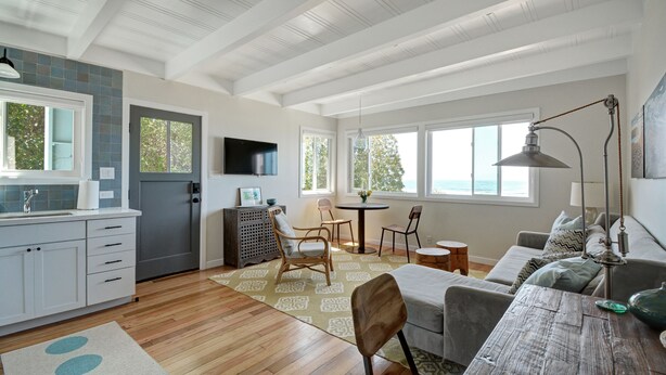 A spacious living area with a wooden floor, a dining table, and a sofa. There is a kitchen area with white cabinets and a sink. Large windows provide natural light and a view of the outdoors. - Oceanfront At Stinson - Relax And Refresh (Stinson Beach)