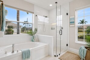 A modern bathroom with a glass-enclosed shower, a freestanding bathtub, and large windows offering views of palm trees and a house.