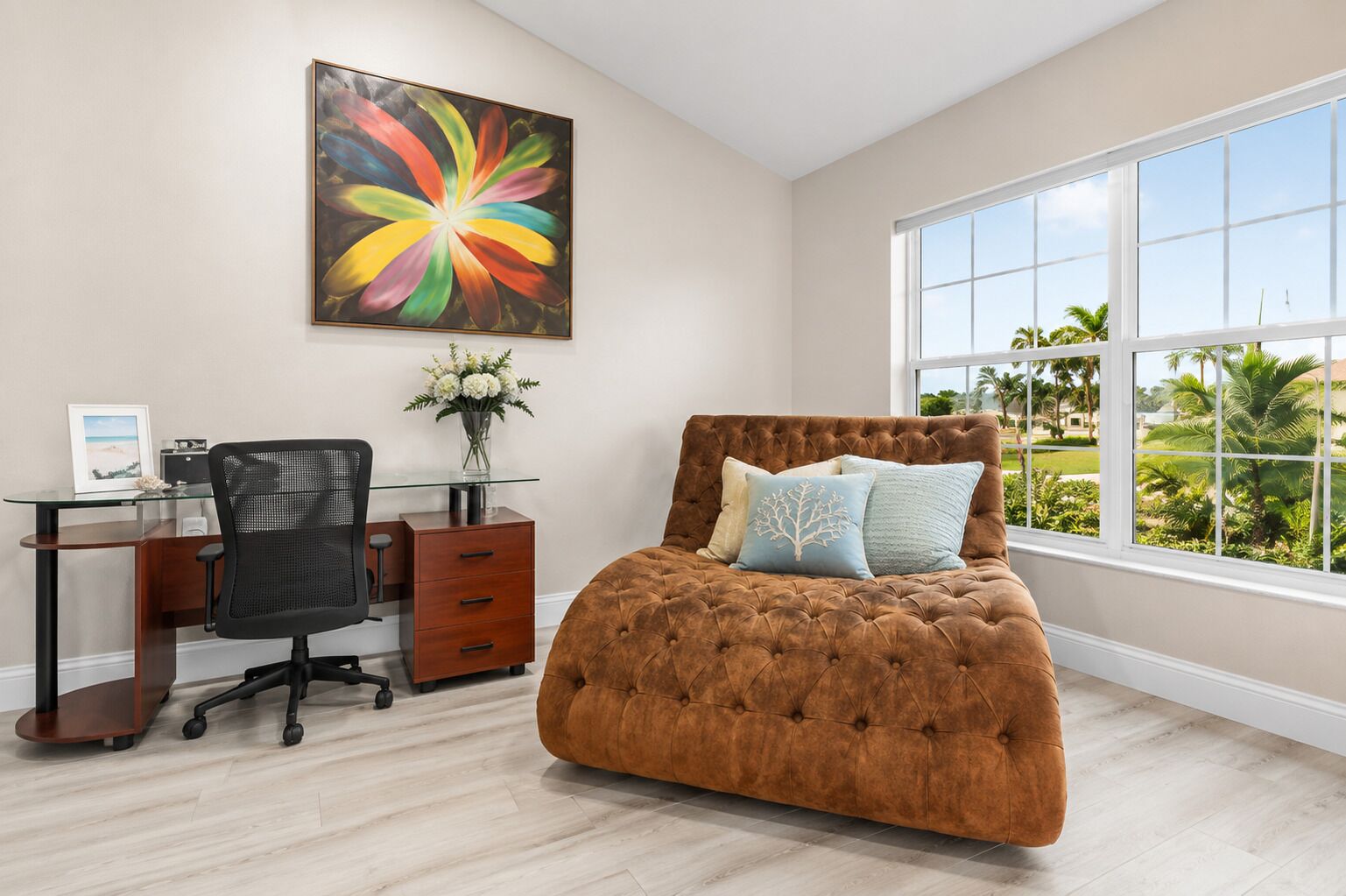 A bedroom with a tufted headboard bed, a glass desk with a chair, a framed picture, and a vase with flowers.