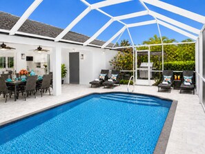 A swimming pool with a white roof structure, a dining area with chairs and a table, and a barbecue grill.