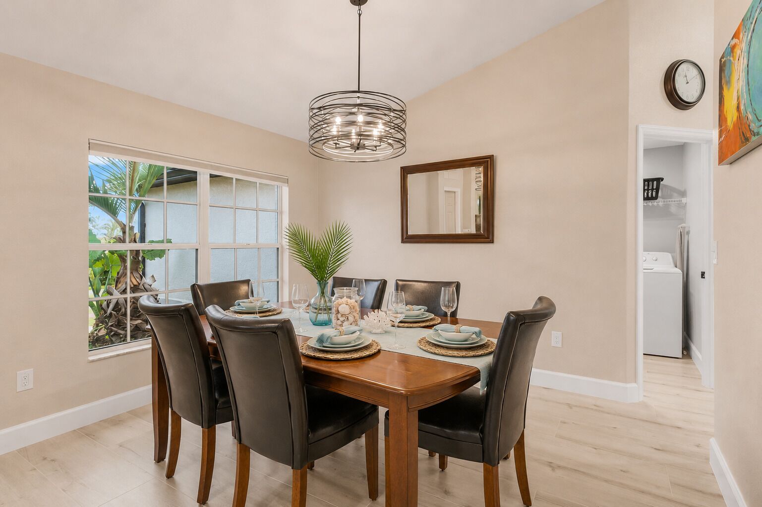 A dining room with a wooden table, black chairs, a hanging light fixture, a window with a view of greenery, and a mirror on the wall.