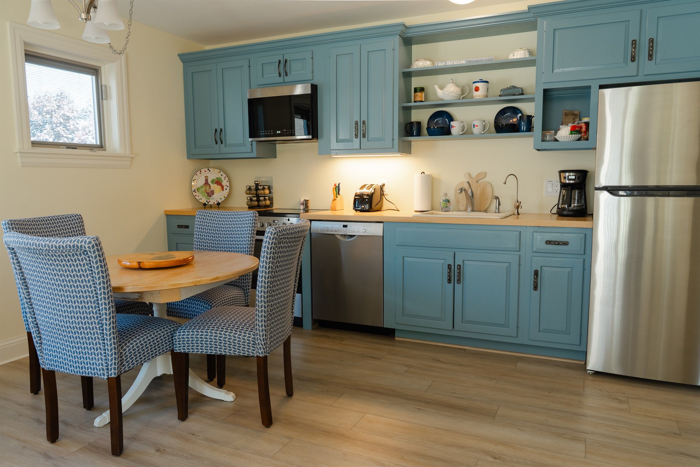 A kitchen with blue cabinets, a stainless steel refrigerator, a dishwasher, and a dining area with patterned chairs.