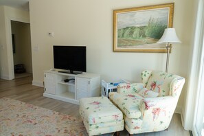 A living room with a flat-screen TV on a white cabinet, a floral armchair, a painting on the wall, and a floor lamp. - Rent Two Suites On A Beautiful Five Acre Estate (Leland)