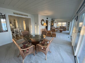A spacious living room with a round glass table surrounded by wicker chairs with floral upholstery. There is a dining area with a white table and chairs, and a kitchen visible in the background.