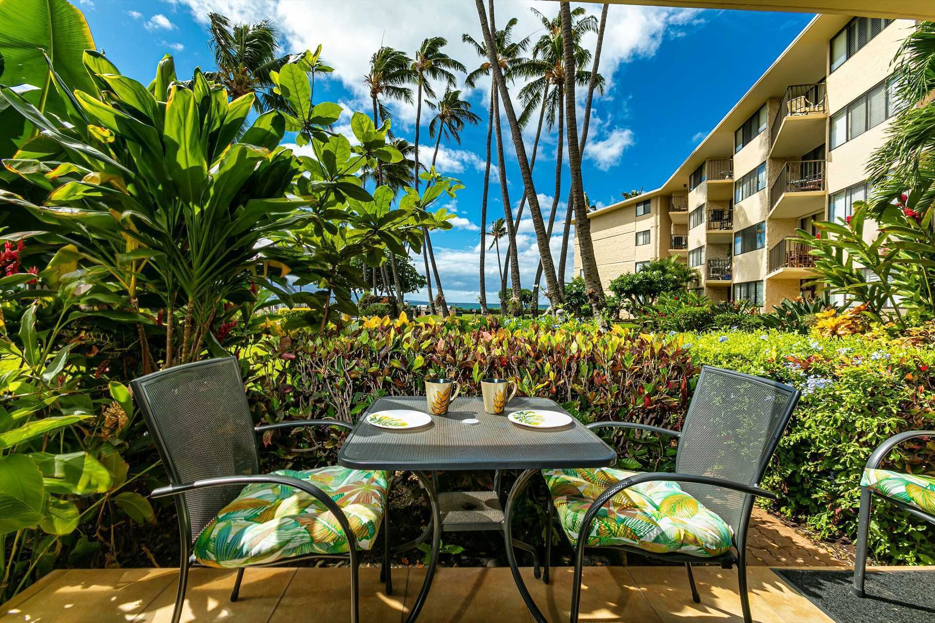 An outdoor seating area with a table and chairs, surrounded by lush tropical plants and palm trees, with a building in the background.