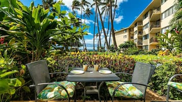 An outdoor seating area with a table and chairs, surrounded by lush tropical plants and palm trees, with a building in the background.