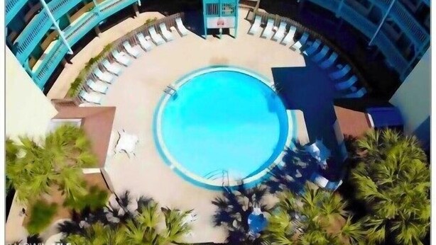 Ein kreisförmiger Pool, umgeben von Liegestühlen und Palmen, mit einem mehrstöckigen Gebäude im Hintergrund.