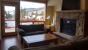A living room with a stone fireplace, a sofa, a coffee table, and a balcony with a view of mountains and houses. - New 3BR/3BA & Loft, Private Hot Tub, Heated Pool, Views, Walk/Bus to River Run (Keystone)