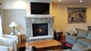 A living room with a stone fireplace, a mounted TV, a sofa, and a painting on the wall. - New 3BR/3BA & Loft, Private Hot Tub, Heated Pool, Views, Walk/Bus to River Run (Keystone)