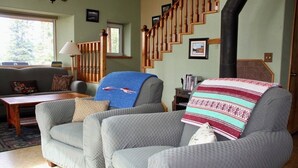 A living room with a grey sofa, a wooden staircase, and a fireplace. - ☆ Denali Sunset House - Wild scenery with creature comforts! (Denali National Park)