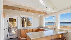 A kitchen with wooden cabinets, a granite countertop, and a large island with bar stools. There are pendant lights, a view of the beach, and a door leading outside.