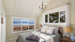 A bedroom with a large bed, two windows with a view of the beach, a chandelier, and a wooden nightstand.