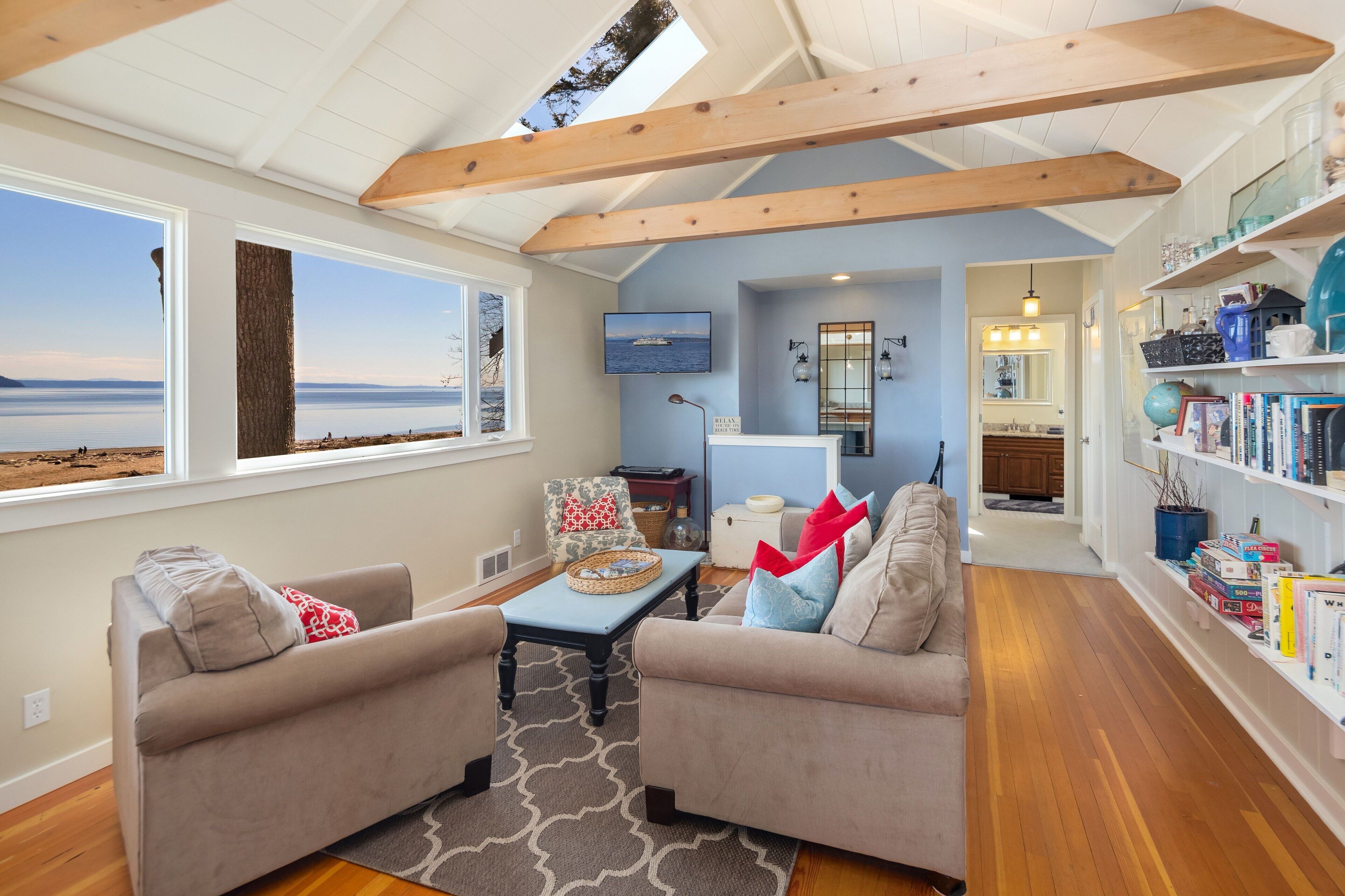 A living room with a view of the sea, a wooden ceiling, a bookshelf, and a cozy seating area.