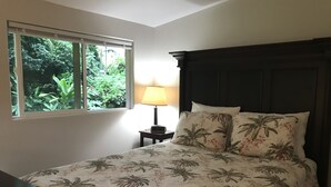 A bedroom with a bed, a nightstand, a lamp, and a window with a view of greenery. - Secluded cottage on Tropical flower farm (Hana)