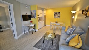 A modern living room with a glass coffee table, a sofa with cushions, a dining area with a table and chairs, and a kitchen with appliances and a TV.