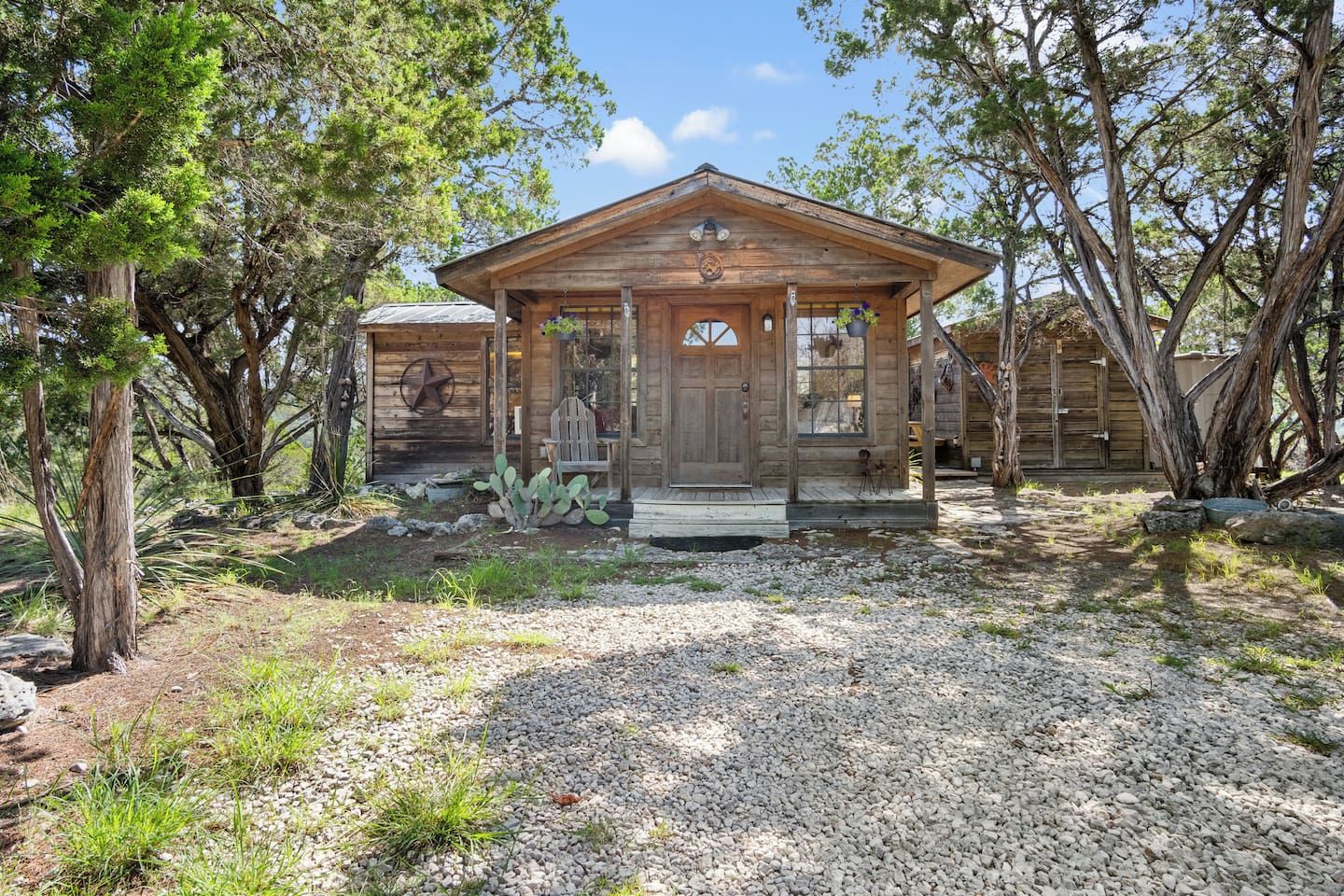 A rustic wooden cabin with a porch, surrounded by trees and a gravel driveway.