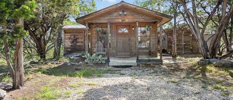A rustic wooden cabin with a porch, surrounded by trees and a gravel driveway.