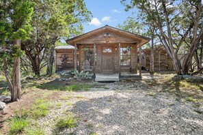 A rustic wooden cabin with a porch, surrounded by trees and a gravel driveway.