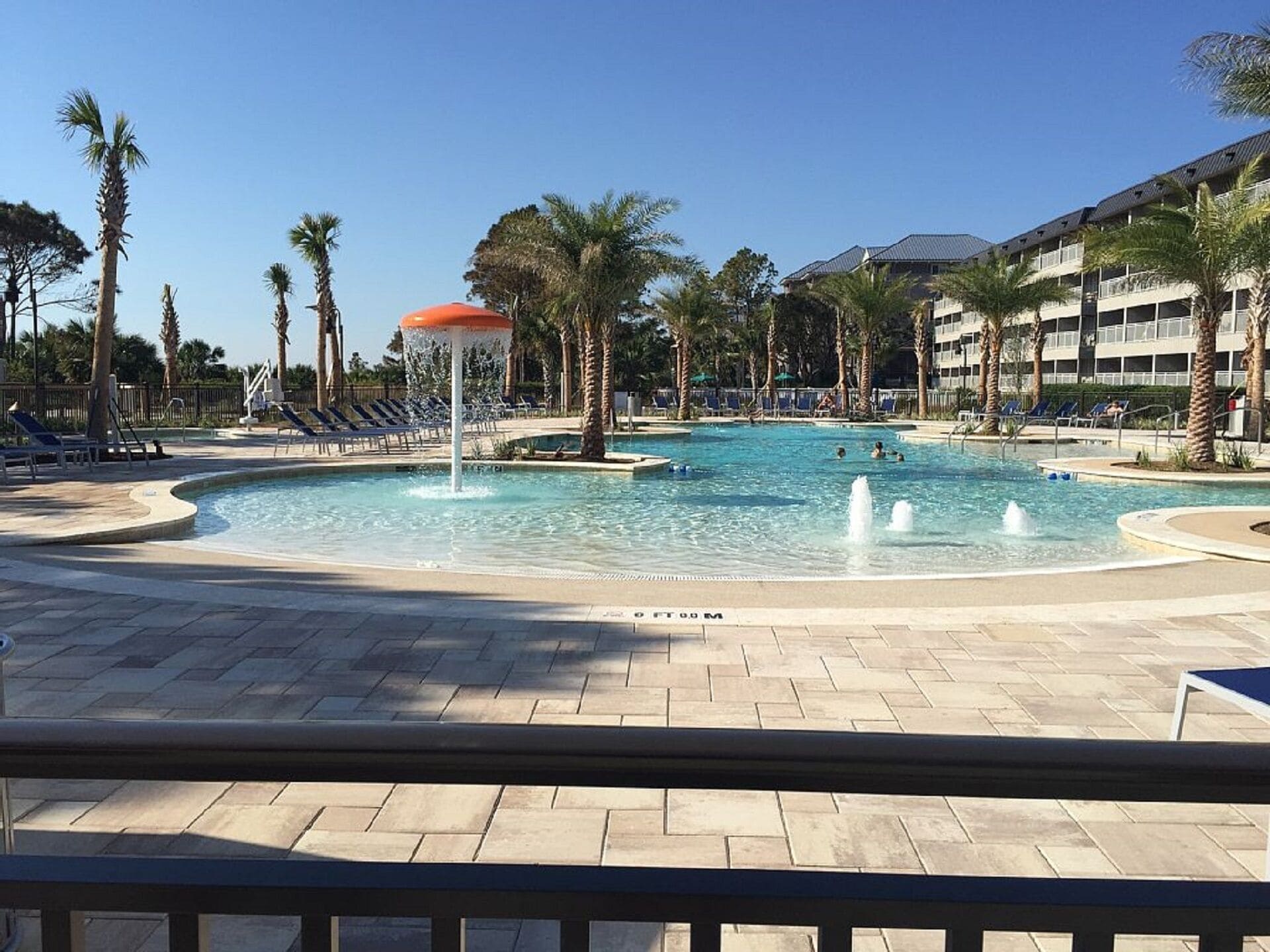 A swimming pool with a water fountain, surrounded by palm trees and lounge chairs.