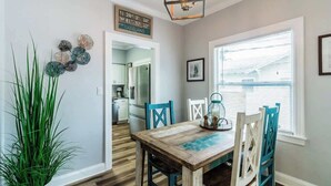Un coin repas avec une table en bois, des chaises bleues, une plante en pot, des éléments décoratifs sur les murs et une fenêtre avec des stores.