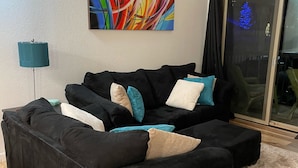 Un salon moderne avec un canapé d’angle noir, un lampadaire turquoise et un tableau abstrait aux couleurs vives accroché au mur.