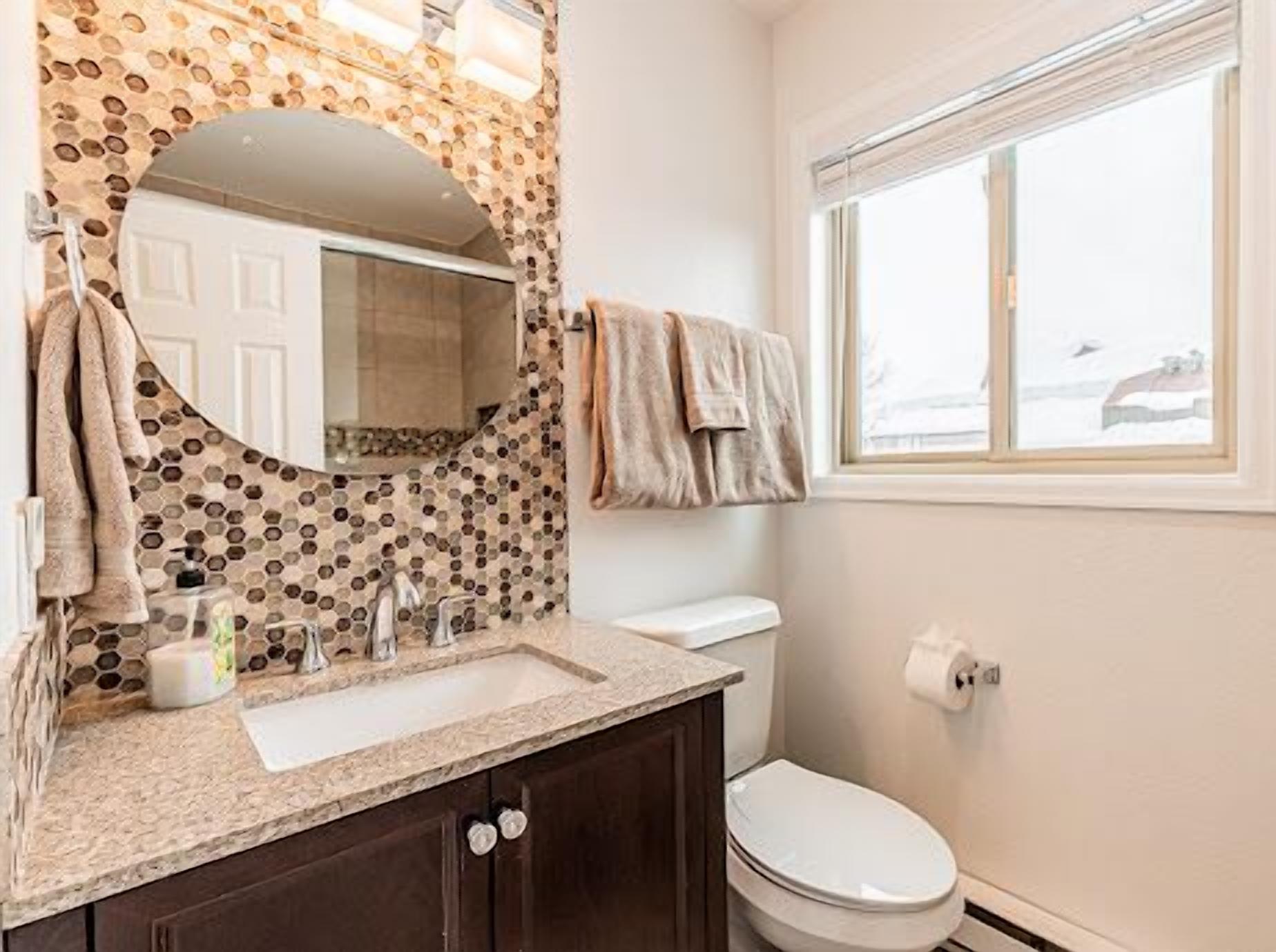 Un baño con salpicadero de mosaico, un espejo redondo, un lavabo con grifo, un inodoro y una ventana con vistas al exterior.