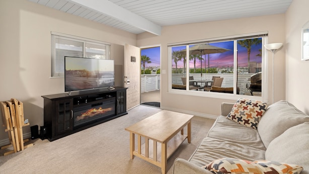 Een woonkamer met een televisie, een open haard en uitzicht op palmbomen en een zonsondergang door grote ramen.