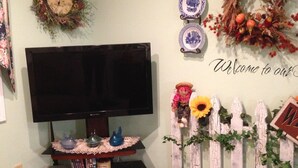 A living room with a flat-screen TV on a stand, a small shelf with decorative items, a teddy bear on a bench, and a wall with a 'Welcome to our home' sign. - Private one bedroom, one bathroom, apartment with private entrance & hot tub (Branson)