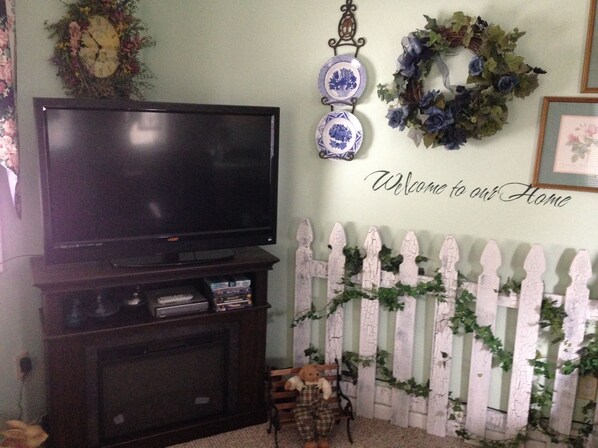 A living room with a flat-screen TV on a stand, a white picket fence, and a welcome sign on the wall. - Private one bedroom, one bathroom, apartment with private entrance & hot tub (Branson)