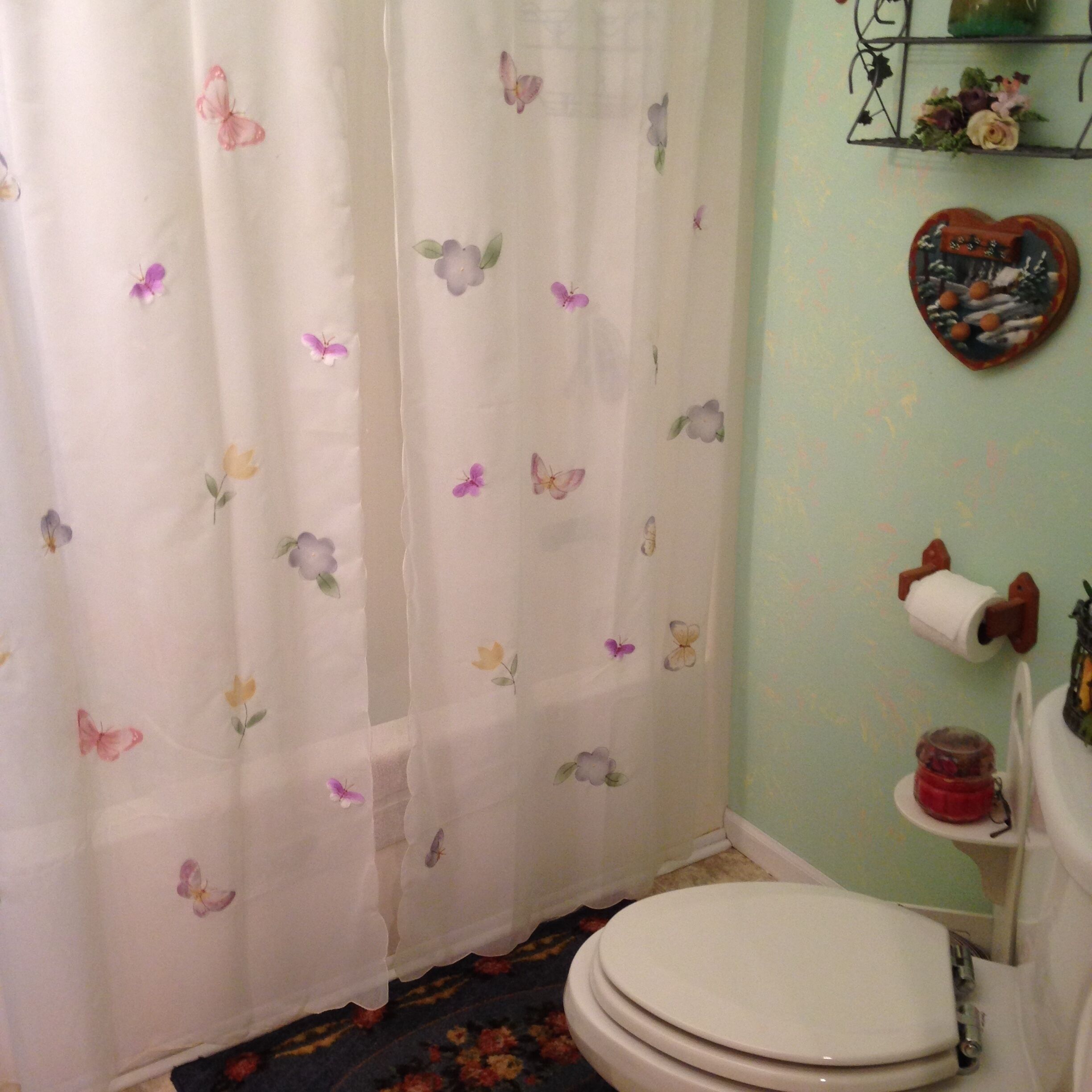A bathroom with a toilet, a toilet paper holder, a heart-shaped decoration, and a floral shower curtain.