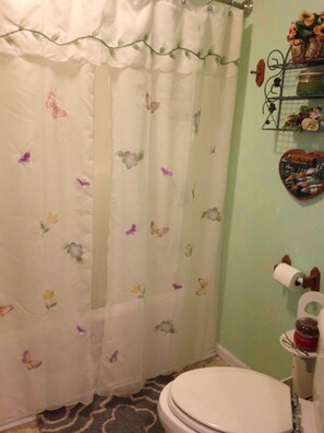 A bathroom with a shower curtain featuring floral and butterfly patterns, a toilet, and a shelf with decorative items. - Private one bedroom, one bathroom, apartment with private entrance & hot tub (Branson)