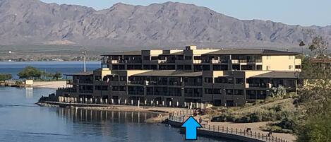 A multi-story hotel with a waterfront view, located near a body of water with mountains in the background.