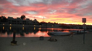 A lakeside scene at sunset with boats docked, a sandy beach, and a building in the background.