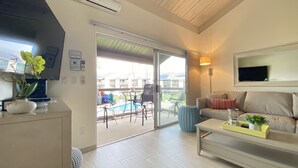 A modern living room with a sofa, coffee table, and a TV mounted on the wall. There is a sliding glass door leading to an outdoor area with a pool and chairs.