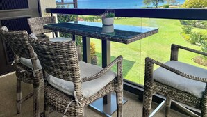 A balcony with wicker chairs and a glass table overlooking a grassy area and the sea.
