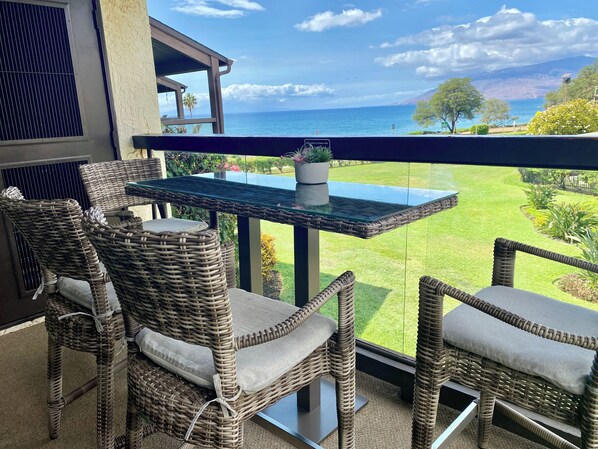 A balcony with wicker chairs and a glass table overlooking a grassy area and the sea.