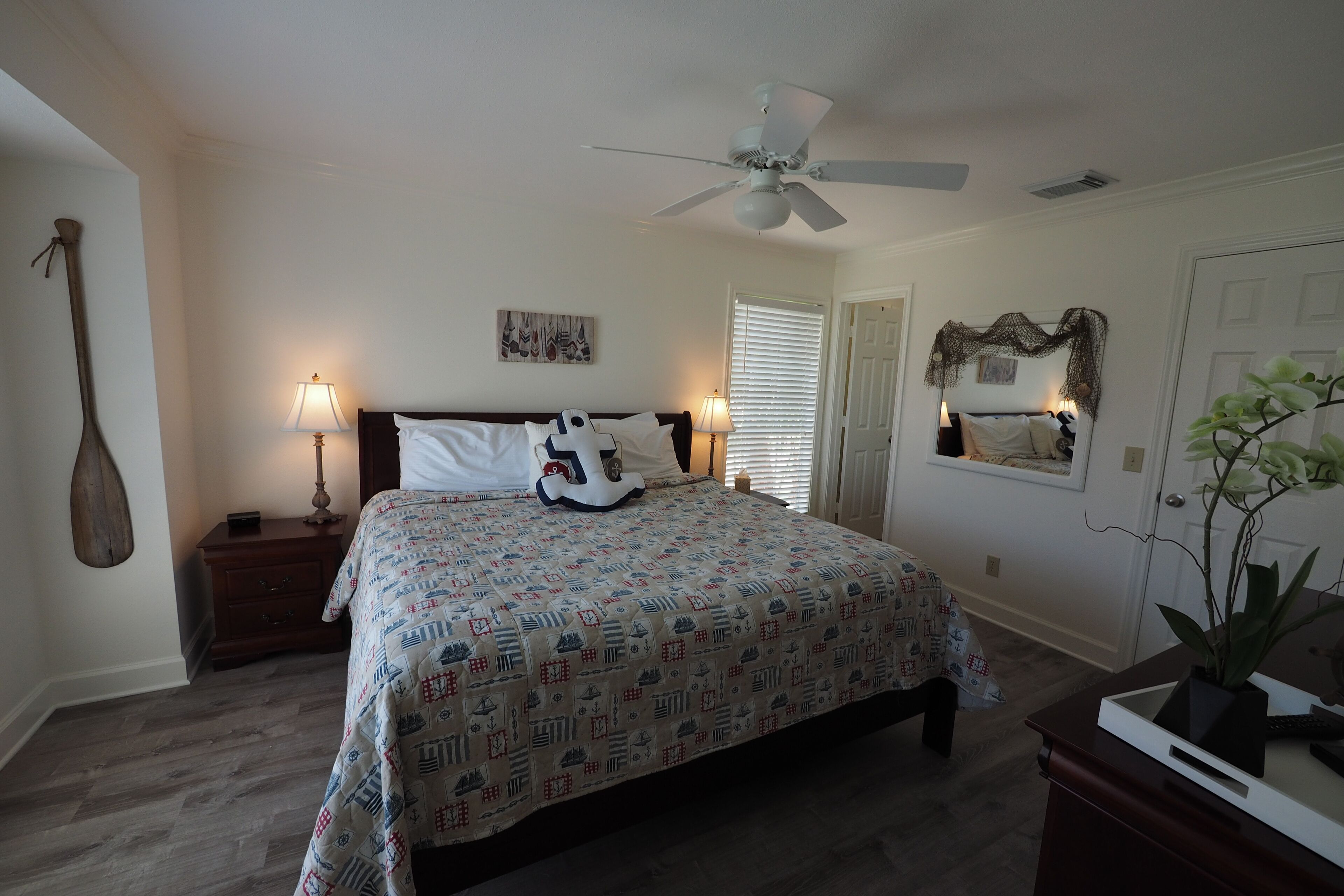 A bedroom with a bed, nightstands, a ceiling fan, a lamp, a door, and a plant.
