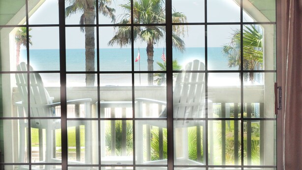 Vista a través de una ventana con un diseño de cuadrícula, mostrando una playa, palmeras y dos sillas blancas.