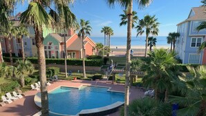 Zona de piscina con sillones reclinables y palmeras, rodeada de edificios con vista a la playa.