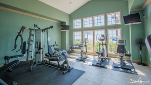 Un gimnasio con diferentes equipos de ejercicio, ventanas amplias y un televisor en la pared.