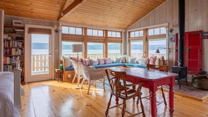 Interior acolhedor de uma cabana com teto de madeira, uma janela grande com vista panorâmica, uma área de refeições com mesa e cadeiras, e uma estante cheia de livros.