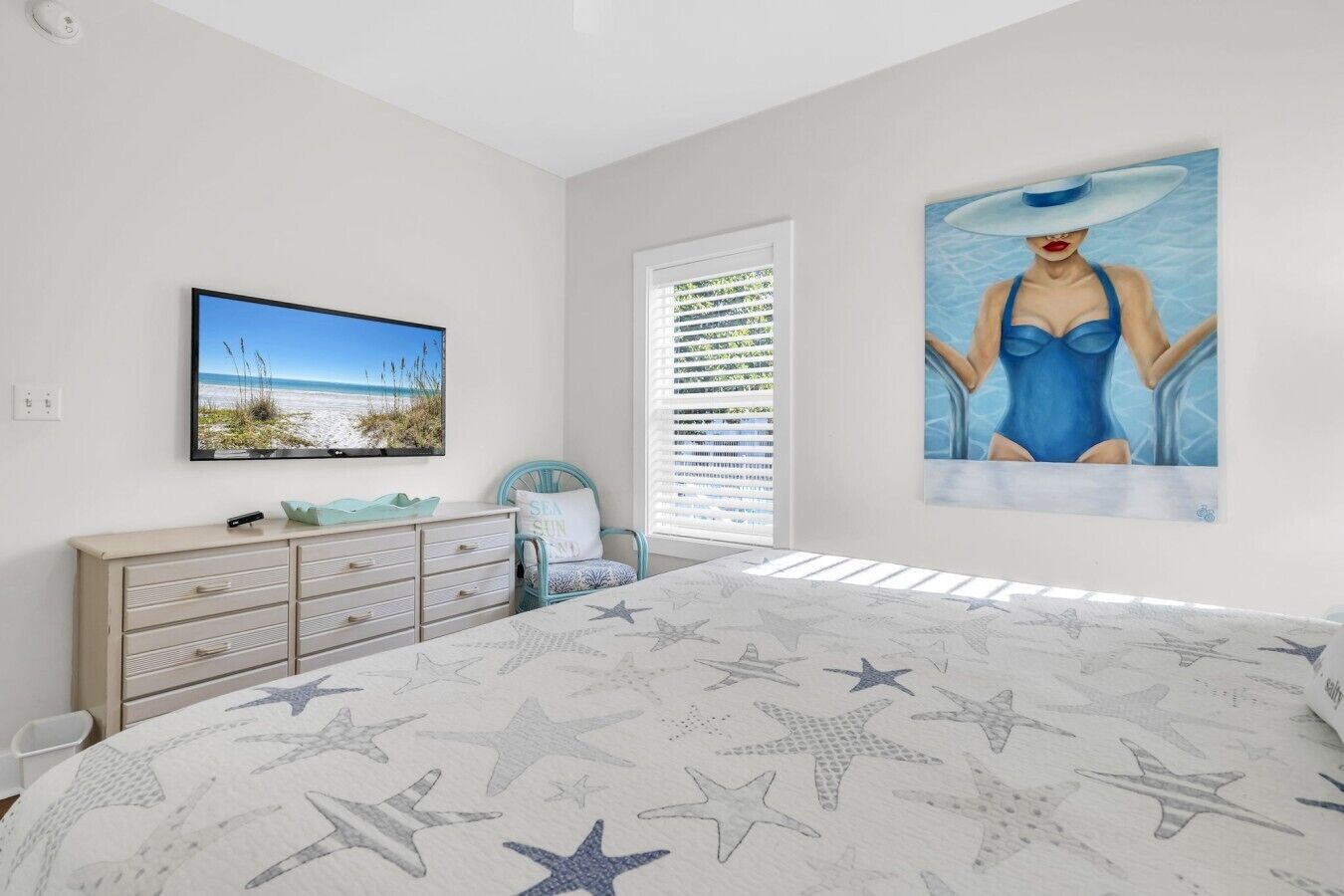 Ein Schlafzimmer mit einem Bettlaken mit Strandmotiv, einem Wandfernseher, einer Kommode und einem Fenster mit Jalousien.