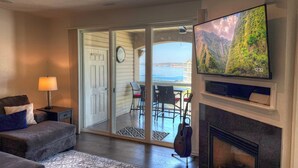 A living room with a sofa, a lamp, a television, a fireplace, and a sliding glass door leading to a balcony with a view of the sea.
