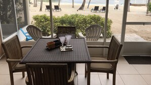 Un espace repas comprenant une table et des chaises, donnant sur une plage bordée de palmiers et offrant une vue sur la mer.