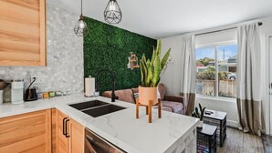 A modern kitchen with a white marble countertop, a black sink, wooden cabinets, and a green wall. There is a dining area with a sofa and a potted plant.