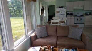 A cozy living room with a brown sofa, a coffee table with books and a remote, and a view of a kitchen and garden through a window. - The Guest Cottage in Paradise. (Northwood)